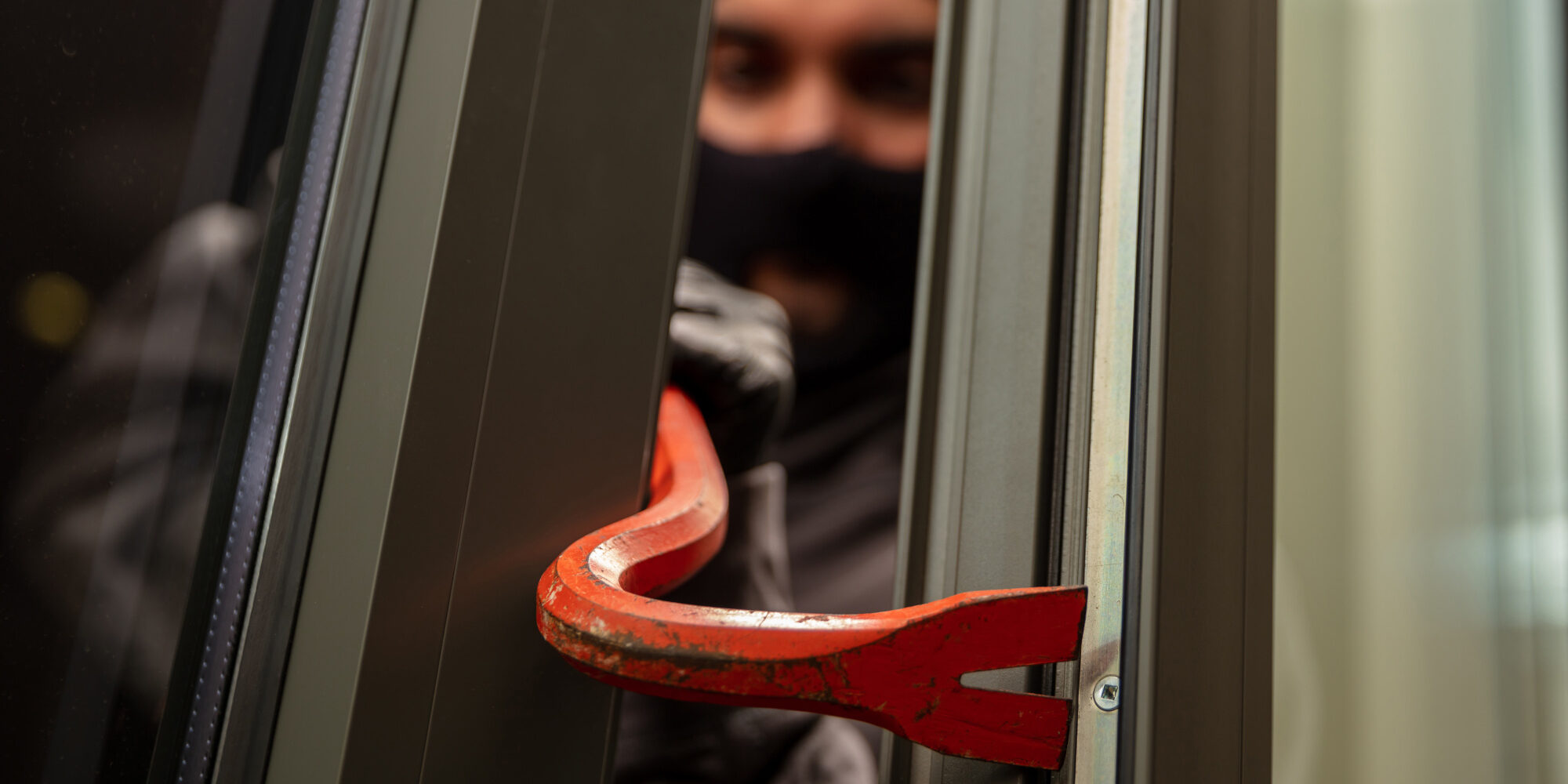 Thief in balaclava holding a crowbar to break a glass window.