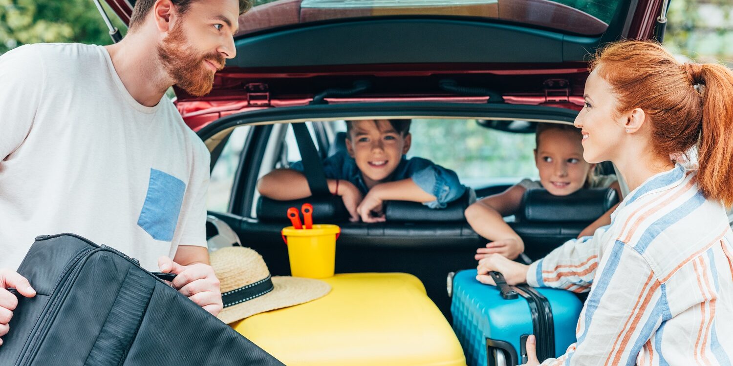 young parents packing luggage in trunk of car with kids on backseats